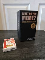 Closed black What Do You Meme? Bigger Better Edition game box standing upright with a closed deck of Victoria playing cards in front on black floor.