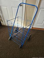 Front angle view of blue metal wire rolling shopping cart showing large open basket, two large rear wheels, and two small front wheels.