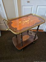 Side view of the bar trolley showing the wood grain, floral inlay on the top shelf, brass rail, and wooden handles.