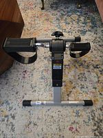 Front view of the mini pedal exerciser showing pedal straps, central resistance knob, and display panel.