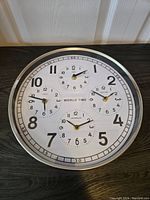 Front view of round world time clock showing main dial and three subdials for different timezones. Clock has metal frame and clear numbering.