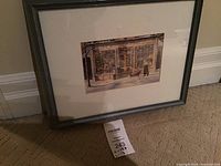 Framed print leaning against a wall showing the winter scene of a storefront with a person and child on a sled, in a grey wood frame.