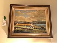 Full view of framed oil painting of Irish countryside with cottages, stone walls, water, and hills under clouds, in wood and gold frame.