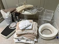 Photo showing assorted bath towels folded in a stack, raised toilet seat, vintage mechanical scale, metal trash cans, white vanity stool, storage baskets, and bath rack on tiled floor.