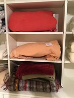 Five folded blankets and throws stacked on shelving unit including a rose pink and peach wool blankets on separate shelves and three throws stacked below.
