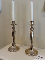Pair of silver plated candlesticks each with a white candle, shown side by side on a white surface.