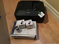 Canon Pixma printer on top of a stack of copy paper, label sheets, and ink cartridge boxes with additional smaller cartridge on top.