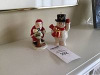 Two Belleek Living Holiday Collection figurines displayed on a white surface: Santa holding a globe, and a snowman holding a present, both with hanging hooks on hats.