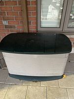 Front view of beige and dark green Rubbermaid plastic deck box outdoors on patio stones with brick wall background.
