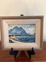 Front view of the framed painting on a black stand, showing the full artwork, matting, and frame.