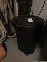 Full view of black Rubbermaid garbage can with lid, placed on garage floor near plumbing pipes and a broom.