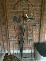 Front view of metal garden trellis with bird and heron designs leaning against a brick wall. Two colorful rooster shapes at the top and black silhouette of a heron among reeds below.