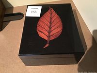 Closed black lacquered cigar humidor box with a large reddish-brown leaf design on top, photographed from above.