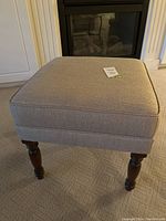 Front view of beige fabric ottoman showing square cushioned top with chevron pattern and wooden legs.