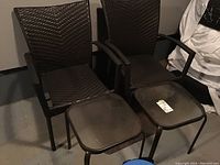 Two brown plastic rattan-style outdoor chairs with armrests and two square brown metal tables with dark glass tops placed indoors on a concrete floor.