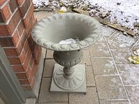 Side view of grey stone-colored cast iron planter next to a brick wall on outdoor paving with light snow visible around.