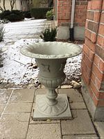 Front view of heavy stone-colored cast iron planter outdoors next to brick wall on paver stones, showing ornamental rim, floral relief accents, rounded stem, and square base.