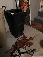 Image showing tall black plastic planter, decorative straw owl on top, and metal tree branch wall hooks with brown oak leaves on the floor