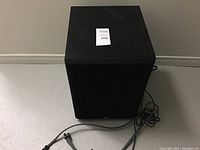 Front view of the black PSB speaker showing cube shape and fabric front grille with lot tag on top.