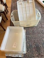 Large clear tote with lid partially removed showing interior, sits on patterned area rug, dining chairs visible in background.