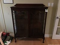 Full view of vintage wooden cabinet with glass front doors and shelving inside, standing on carved legs.