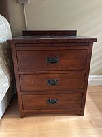 Front view of wooden night table showing three drawers with dark metal pulls and knob, dark wood grain finish.