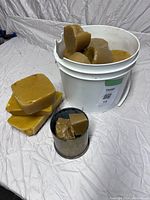 Full view showing white bucket filled with differently shaped beeswax blocks alongside several beeswax blocks outside on cloth.