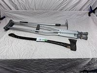 Full view showing a pair of aluminum crutches, a manual axe, and an extendable garden weeder tool laid flat on a white crinkled cloth.
