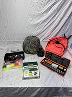 Overview photo showing the entire lot including camouflage firearm cleaning pad, fishing lure box, firearm cleaning kit case, and bright orange backpack laid out on a white surface.