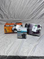 Three boxed items on white fabric backdrop: Black & Decker angle grinder box, Intex electric air pump box, and purple/white iron box