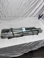 Folded metal frame military cot held together with green fabric strap, lying on white fabric surface.