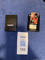 Photo showing Zippo lighter inside black Zippo branded plastic case alongside its black outer box with Zippo logo.