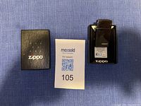 Photo of Zippo lighter with its black box and a support card on a blue fabric surface.