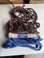 View of the three ropes: a twisted white cotton rope packaged, a thick black and orange braided rope, and a thinner blue braided rope.