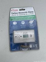Front view of Kidde carbon monoxide alarm in sealed original packaging, showing model number, digital display, and code of conduct in English and Spanish.