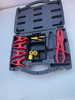 Open black plastic carrying case showing content layout with red clamps, yellow connectors, and a red plier tool