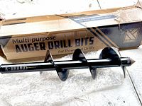 Photo of one black auger drill bit with spiral blade and pointed tip lying in front of its multi-purpose auger drill bits cardboard box.