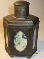 Front and side images showing the dark pewter hexagonal tea caddy with ornate embossed borders framing hand-painted ceramic panels depicting Chinese figures in traditional robes and settings.