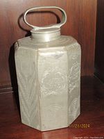 Front view of the antique pewter tea caddy showing engraved floral patterns and oval handle on the lid.