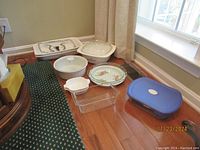 Overview of multiple bakeware items including casserole dishes, pie dishes, glass baking pan and a plastic storage container placed on a wooden floor near a carpet and window.