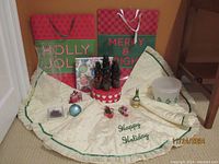 Two oversized Christmas gift bags, cream tree skirt with embroidered Happy Holiday, miniature trees, and several ornaments on floor.