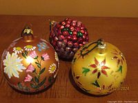 Three Christopher Radko Christmas ornaments displayed on a brown surface, showing each ornament clearly: floral English Garden, gold poinsettia, and cranberry cluster