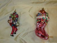 Photo of two Christopher Radko ornaments on a beige fabric background showing a Bell Hop carrying gifts and a red stocking filled with gifts and candy cane.
