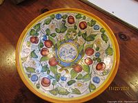 Top-down photo of a large round decorative wall hanging dish with colorful hand-painted fruit and floral patterns and a central emblem.