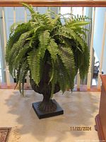 Faux fern plant with green leaves in resin urn, standing on a carpeted floor near railing.
