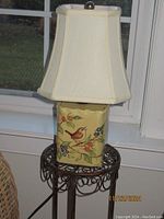 Photo of lamp with hand painted ceramic base featuring bird and berry branch, cream fabric lampshade, placed on metal stand with scroll detail
