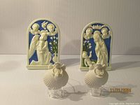 Photo showing two Halcyon Days terracotta relief sculptures with religious scenes, both with blue backgrounds and ivory figures, along with two small cream-colored seashell ornaments in front.