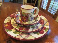 Stacked view showing a mug, bowl, salad plate, and dinner plate with colorful hand-painted floral and fruit design and burgundy rims on a wooden table.