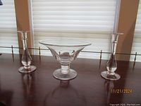 Three glass art pieces on dark wooden surface, white blinds background: footed pedestal compote bowl in center flanked by candle holders.