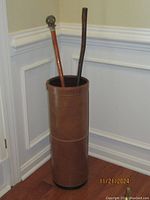 Photo of Stetson leather umbrella stand holding two walking sticks in a corner of a room with wood flooring and white wainscoting.